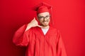 Young redhead man wearing red graduation cap and ceremony robe smiling doing phone gesture with hand and fingers like talking on Royalty Free Stock Photo