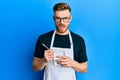 Young redhead man wearing waiter apron taking order in shock face, looking skeptical and sarcastic, surprised with open mouth