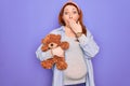 Young redhead pregnant woman expecting baby holding teddy bear over purple background cover mouth with hand shocked with shame for Royalty Free Stock Photo