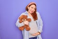 Young redhead pregnant woman expecting baby holding teddy bear over purple background with a happy face standing and smiling with Royalty Free Stock Photo