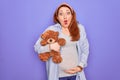 Young redhead pregnant woman expecting baby holding teddy bear over purple background scared in shock with a surprise face, afraid Royalty Free Stock Photo