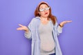 Young redhead pregnant woman expecting baby wearing funny thug life sunglasses clueless and confused expression with arms and Royalty Free Stock Photo