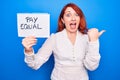 Young redhead woman asking for equality economy holding paper with pay equal message pointing thumb up to the side smiling happy