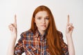 Young redhead woman biting lip with complicated look, pointing fingers up and staring worried at camera, standing over