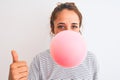 Young redhead woman chewing gum and blowing hair bubble over white isolated background happy with big smile doing ok sign, thumb