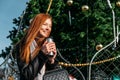 Young redhead woman drinking coffee wandering near Christmas tree in the streets on winter city. Happy young smiling Royalty Free Stock Photo