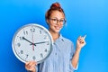 Young redhead woman holding big clock smiling happy pointing with hand and finger to the side Royalty Free Stock Photo