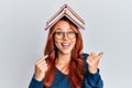 Young redhead woman holding books over head pointing thumb up to the side smiling happy with open mouth Royalty Free Stock Photo