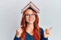 Young redhead woman holding books over head smiling happy pointing with hand and finger to the side Royalty Free Stock Photo