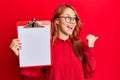 Young redhead woman holding clipboard with blank space pointing thumb up to the side smiling happy with open mouth Royalty Free Stock Photo