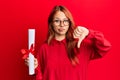 Young redhead woman holding graduate degree diploma with angry face, negative sign showing dislike with thumbs down, rejection Royalty Free Stock Photo