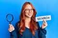 Young redhead woman holding magnifying glass and search word winking looking at the camera with sexy expression, cheerful and