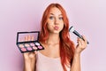 Young redhead woman holding makeup and brush puffing cheeks with funny face