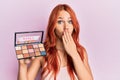 Young redhead woman holding makeup nudes covering mouth with hand, shocked and afraid for mistake