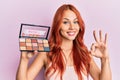 Young redhead woman holding makeup nudes doing ok sign with fingers, smiling friendly gesturing excellent symbol