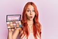 Young redhead woman holding makeup nudes scared and amazed with open mouth for surprise, disbelief face