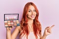 Young redhead woman holding makeup nudes smiling happy pointing with hand and finger to the side