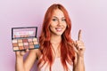 Young redhead woman holding makeup nudes smiling with an idea or question pointing finger with happy face, number one