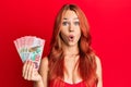 Young redhead woman holding 100 new zealand dollars banknote scared and amazed with open mouth for surprise, disbelief face Royalty Free Stock Photo