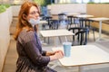 Young redhead woman with a medical mask removed sits in an empty cafe after quarantine. Deserted restaurant after the completion