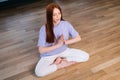 Young redhead woman meditating practicing breathing yoga exercise, sitting on floor near window in lotus position. Royalty Free Stock Photo