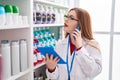 Young redhead woman pharmacist talking on smartphone using touchpad at pharmacy