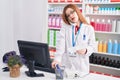 Young redhead woman pharmacist talking on telephone using computer at pharmacy