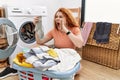 Young redhead woman putting dirty laundry into washing machine shouting and screaming loud to side with hand on mouth