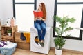 Young redhead woman reading book waiting for washing machine at laundry room Royalty Free Stock Photo