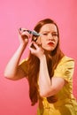 Young redhead woman with red lips doing her eyes makeup, applying eyeliner with knife against pink background Royalty Free Stock Photo