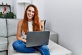 Young redhead woman using laptop at home pointing thumb up to the side smiling happy with open mouth Royalty Free Stock Photo