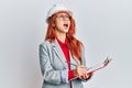 Young redhead woman wearing architect hardhat angry and mad screaming frustrated and furious, shouting with anger looking up Royalty Free Stock Photo