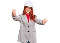 Young redhead woman wearing architect hardhat looking at the camera smiling with open arms for hug Royalty Free Stock Photo