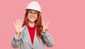 Young redhead woman wearing architect hardhat showing and pointing up with fingers number seven while smiling confident and happy Royalty Free Stock Photo