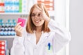 Young redhead woman working at pharmacy drugstore holding condom smiling happy doing ok sign with hand on eye looking through
