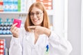 Young redhead woman working at pharmacy drugstore holding condom smiling happy pointing with hand and finger Royalty Free Stock Photo