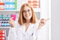 Young redhead woman working at pharmacy drugstore holding condom smiling happy pointing with hand and finger to the side