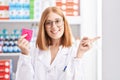 Young redhead woman working at pharmacy drugstore holding condom smiling happy pointing with hand and finger to the side Royalty Free Stock Photo