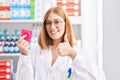 Young redhead woman working at pharmacy drugstore holding condom smiling happy and positive, thumb up doing excellent and approval