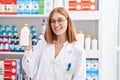 Young redhead woman working at pharmacy drugstore holding sun screen looking positive and happy standing and smiling with a Royalty Free Stock Photo