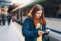 Young redhhead woman with backpack checking her ticket and using smartphone on background train. Railroad transport Royalty Free Stock Photo