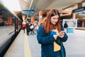 Young redhhead woman waiting on station platform with backpack on background electric train using smart phone. Railroad Royalty Free Stock Photo