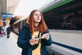 Young redhhead woman waiting on station platform with backpack on background electric train using smart phone. Railroad Royalty Free Stock Photo