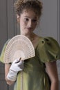 A young Regency woman wearing a green shot silk dress and standing in front of a white paneled wall and holding a fan