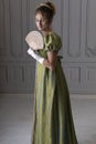 A young Regency woman wearing a green shot silk dress and standing in front of a white paneled wall and holding a fan