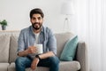 Young relaxed arab guy sitting on couch and drinking tea or coffee, resting in living room interior, free space