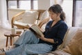 Young relaxed asian woman in casual outfit reading book and drinking tea while relaxing on sofa at home Royalty Free Stock Photo