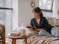 Young relaxed asian woman in casual outfit reading book and drinking tea while relaxing on sofa at home Royalty Free Stock Photo