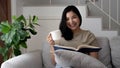 Young relaxed and happy asian woman in casual outfit reading book and drinking tea while relaxing on sofa in cozy living Royalty Free Stock Photo