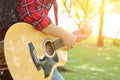 Young relaxed man in red shirt holding an acoustic guitar and playing music at the park outdoors with sunshine filters background.